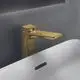 Armatur in gebürstetem Gold von Ideal Standard, mit flacher Auslaufdüse und modernem Design, montiert über einem weißen Waschbecken in einem nüchternen, grauen Umfeld.